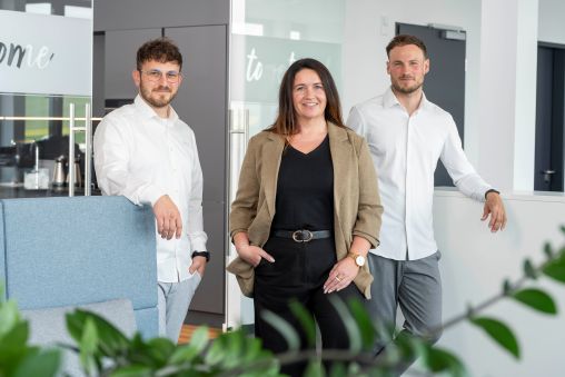 Das Digital-Sales-Team, bestehend aus drei Personen, stehen in einem modernen Büro: zwei Männer in weißen Hemden und eine Frau in einem beigen Blazer.